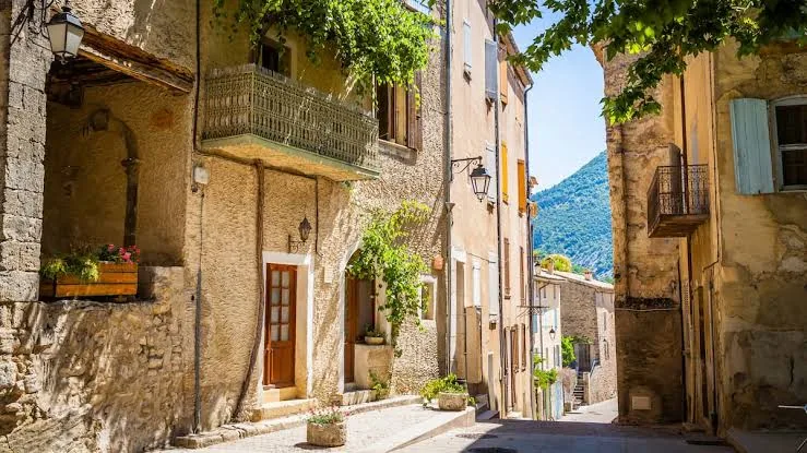 Ruelle en pente bordée de maisons en pierre, de balcons et de plantes, avec les montagnes au loin.