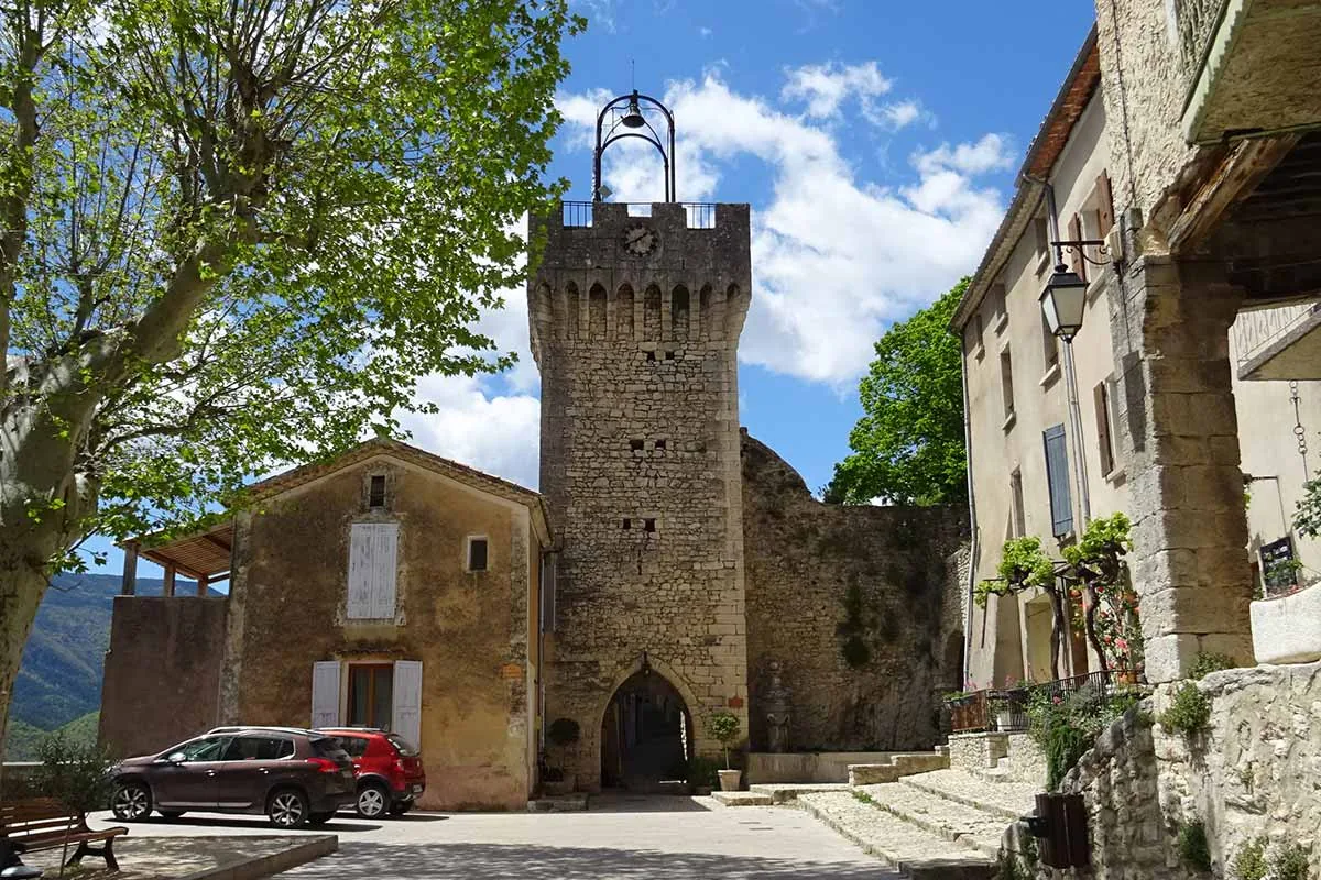 Tour en pierre du beffroi de Montbrun-les-Bains sur une place bordée d’arbres et de maisons.